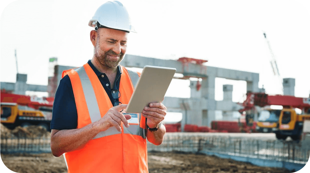 Construction manager in high vis vest and hardhat stands in front of construction site, holding a tablet.